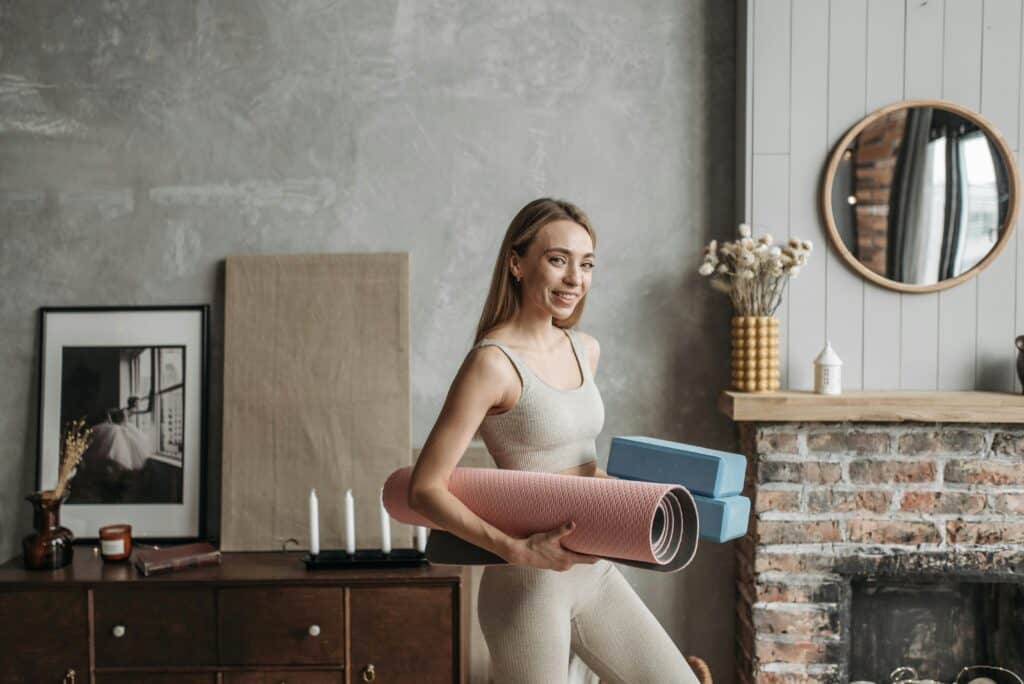 A smiling woman in activewear holding a yoga mat and blocks in a stylish home interior.