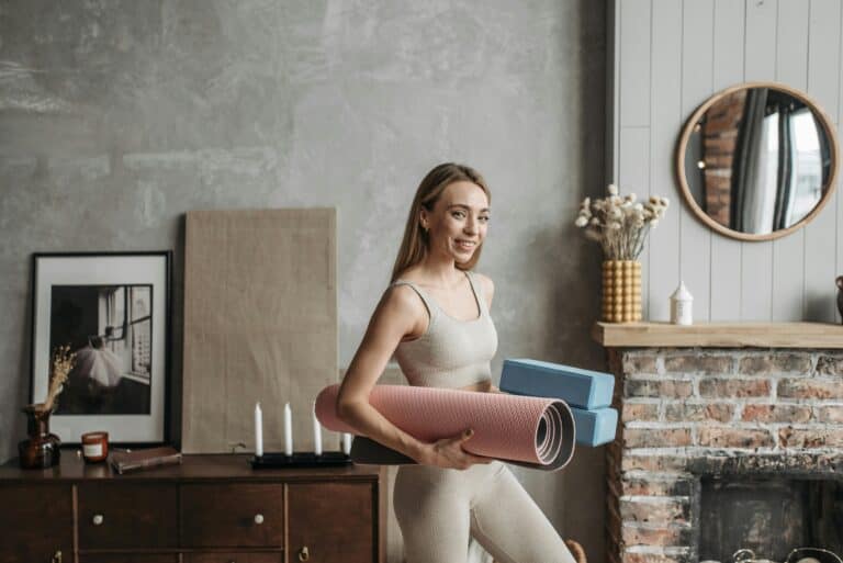 A smiling woman in activewear holding a yoga mat and blocks in a stylish home interior.